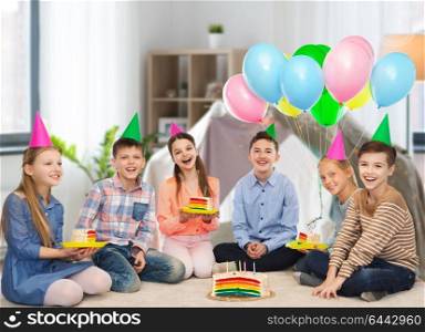 childhood, holidays, celebration, friendship and people concept - happy smiling children in party hats with birthday cake and balloons at home over kids room and tepee background. happy children in party hats with birthday cake