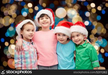 childhood, holidays, friendship and people concept - group of happy smiling little children in santa hats hugging over christmas lights background. happy little children in santa hats hugging