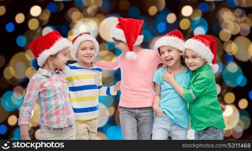 childhood, holidays, friendship and people concept - group of happy smiling little children in santa hats hugging over christmas lights background. happy little children in santa hats hugging