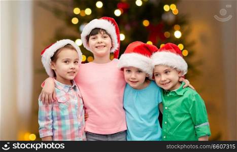 childhood, holidays, friendship and people concept - group of happy smiling little children in santa hats hugging over christmas tree lights background. happy children in santa hats hugging on christmas