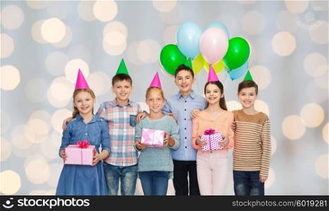 childhood, holidays, friendship and people concept - happy smiling children in party hats with gifts and balloons on birthday over lights background