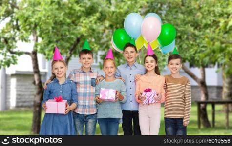 childhood, holidays, friendship and people concept - happy smiling children in party hats with gifts and balloons on birthday over private house backyard background