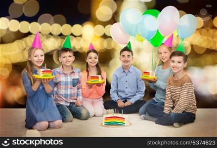 childhood, holidays, friendship and people concept - happy smiling children in party hats with rainbow birthday cake and balloons over festive lights background. happy children in party hats with birthday cake