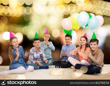 childhood, holidays, friendship and people concept - happy smiling children in party hats with birthday cake and balloons over festive lights background. happy children in party hats with birthday cake