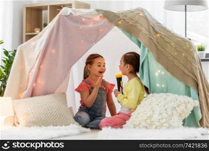 childhood, hygge and friendship concept - happy girls playing with torch light in kids tent at home. girls playing with torch in kids tent at home