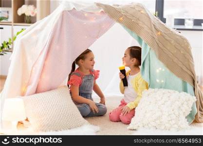 childhood, hygge and friendship concept - little girls with torch light having fun in kids tent or teepee at home. little girls with torch light in kids tent at home