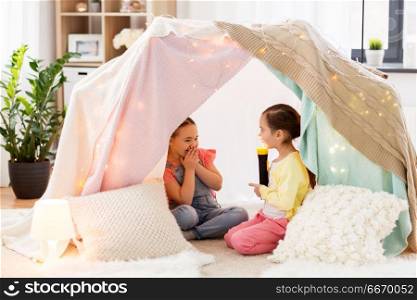 childhood, hygge and friendship concept - little girls with torch light having fun in kids tent or teepee at home. little girls with torch light in kids tent at home. little girls with torch light in kids tent at home