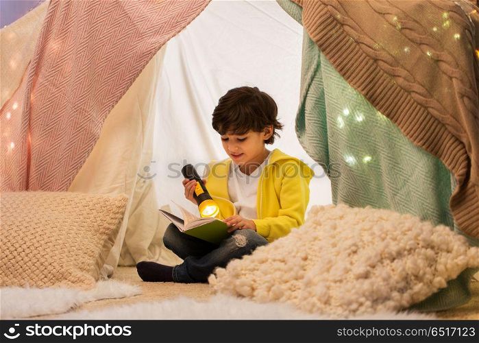 childhood, hygge and people concept - happy boy reading book with torch light in kids tent or teepee at home. happy boy reading book in kids tent at home. happy boy reading book in kids tent at home