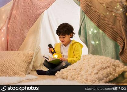 childhood, hygge and people concept - happy boy reading book with torch light in kids tent or teepee at home. happy boy reading book in kids tent at home. happy boy reading book in kids tent at home