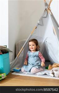 childhood, hygge and people concept - happy little baby girl playing in kid’s tent or teepee at home. baby girl playing in kids tent or teepee at home