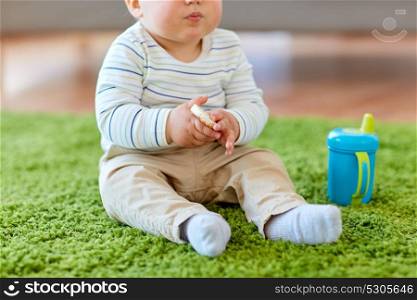 childhood, kids and people concept - baby boy sitting on floor with sippy cup and eating rice cracker at home. baby boy on floor and eating rice cracker at home