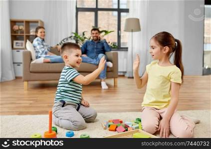 childhood, leisure and family concept - brother and sister playing with wooden toy blocks and making high five gesture at home. brother and sister playing toy blocks at home