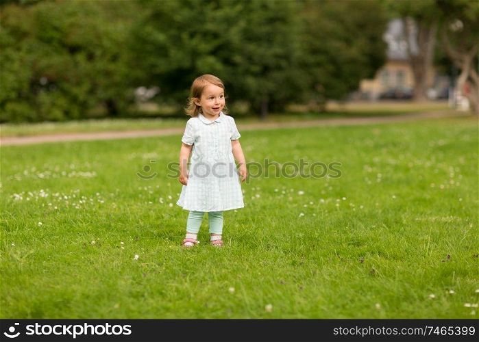 childhood, leisure and people concept - happy baby girl on green meadow at summer park. happy baby girl at summer park