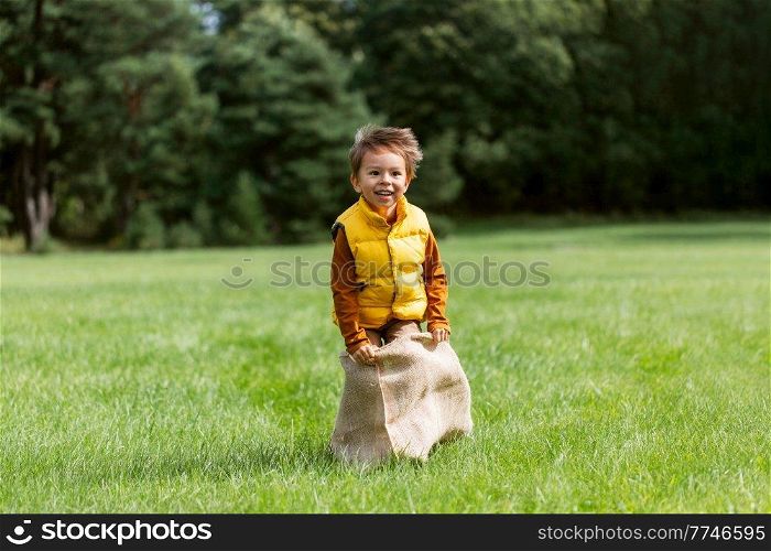 childhood, leisure and people concept - happy little boy playing bag jumping game at park. happy boy playing bag jumping game at park