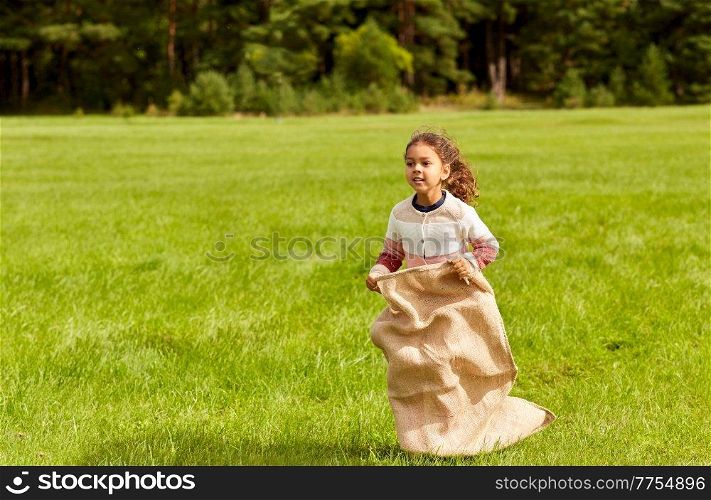 childhood, leisure and people concept - happy little girl playing bag jumping game at park. happy little girl playing bag jumping game at park