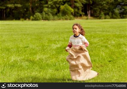 childhood, leisure and people concept - happy little girl playing bag jumping game at park. happy little girl playing bag jumping game at park