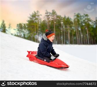 childhood, leisure and season concept - happy little boy sliding on sled down snow hill outdoors in winter over forest or park background. happy boy sliding on sled down snow hill in winter