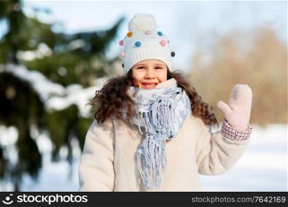 childhood, leisure and season concept - portrait of happy little girl in winter clothes waving hand at park. happy little girl waving hand outdoors in winter
