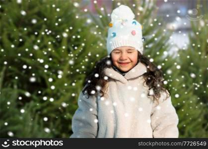 childhood, leisure and season concept - portrait of happy little girl in winter clothes at christmas tree market. happy little girl in winter clothes over snow