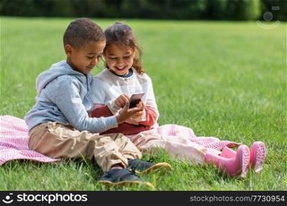 childhood, leisure and technology concept - happy boy and girl with smartphone sitting on blanket at park. kids with smartphone sitting on blanket at park