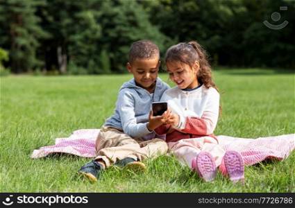 childhood, leisure and technology concept - happy children with smartphone sitting on blanket outdoors. kids with smartphone sitting on blanket outdoors