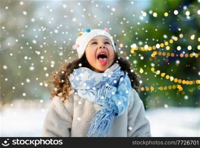 childhood, leisure and winter holidays concept - happy little girl having fun and catching snow by her tongue at winter park. happy girl catching snow by her tongue in winter