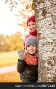 childhood, leisure, friendship, gesture and people concept - happy little girl and boy hiding behind birch tree trunk and waving hand outdoors