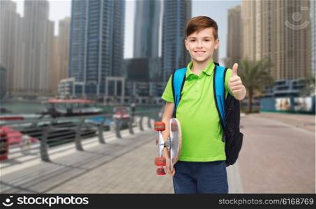 childhood, leisure, gesture, school and people concept - happy smiling student boy with backpack and skateboard showing thumbs up over dubai city street background