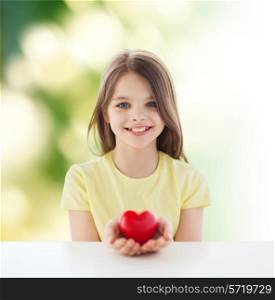 childhood, love, charity, ecology and people concept - beautiful little girl sitting at table and holding red heart over green background