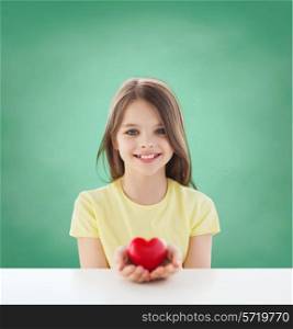childhood, love, charity, education and people concept - smiling little girl sitting and holding red heart over green blackboard background