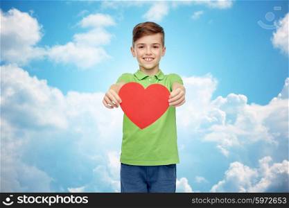 childhood, love, charity, health care and people concept - happy smiling boy in green polo t-shirt holding blank red heart shape over blue sky and clouds background