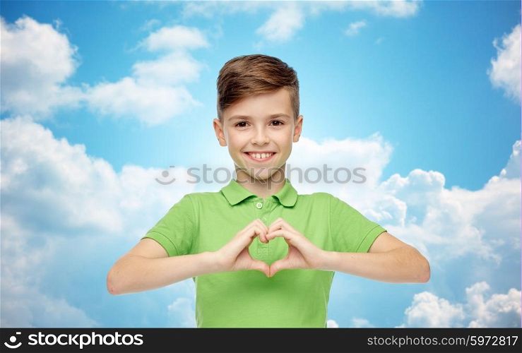 childhood, love, charity, health care and people concept - happy smiling boy in green polo t-shirt showing heart hand sign over blue sky and clouds background