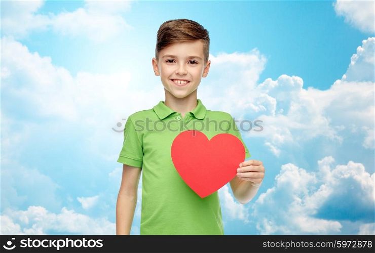 childhood, love, charity, health care and people concept - happy smiling boy in green polo t-shirt holding blank red heart shape over blue sky and clouds background