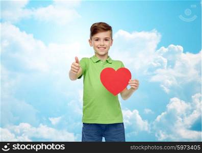 childhood, love, charity, health care and people concept - happy smiling boy in green polo t-shirt holding blank red heart shape and showing thumbs up over blue sky and clouds background