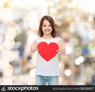 childhood, love, charity, holidays and people concept - smiling little girl sitting with red heart cutout over sparkling background