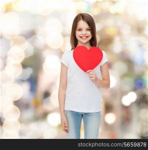 childhood, love, charity, holidays and people concept - smiling little girl sitting with red heart cutout over sparkling background