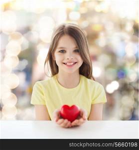 childhood, love, charity, holidays and people concept - smiling little girl sitting and holding red heart over sparkling background