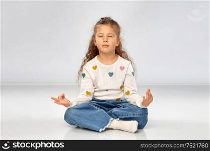 childhood, mindfulness and meditation concept - beautiful little girl in yoga lotus pose and meditating over grey background. little girl in yoga lotus pose and meditating