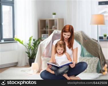 childhood, parenting and relationship concept - happy mother with adorable little daughter reading book at home over kids room and tepee background. happy mother with little daughter reading book