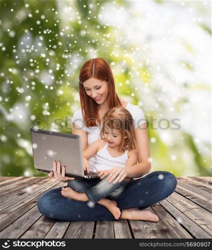 childhood, parenting, people and technology concept - happy mother with little girl with laptop computer over wooden floor and green plants background
