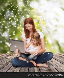 childhood, parenting, people and technology concept - happy mother with little girl with laptop computer over wooden floor and green plants background
