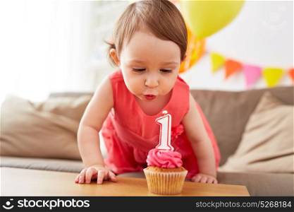 childhood, people and celebration concept - happy baby girl blowing to candle with number 1 on cupcake on birthday party at home. girl blowing to candle on cupcake at birthday