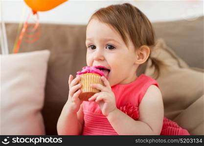 childhood, people and celebration concept - happy baby girl eating cupcake on birthday party at home. happy baby girl eating cupcake on birthday party