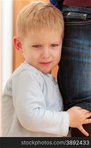 Childhood. Portrait of unhappy angry crying blond boy child kid son hugging leg of his mother at home. Emotions and feelings.