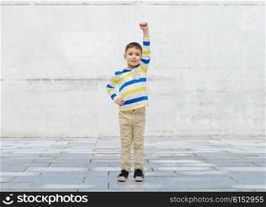 childhood, power, gesture and people concept - happy smiling little boy with raised hand over urban concrete background