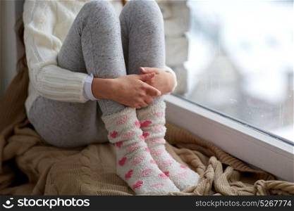 childhood, sadness and people concept - close up of girl in sweater sitting on sill at home window in winter. girl sitting on sill at home window in winter