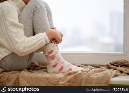 childhood, sadness and people concept - close up of girl in sweater sitting on sill at home window in winter. girl sitting on sill at home window in winter