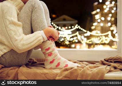 childhood, sadness and people concept - close up of girl in sweater sitting on sill at home window over christmas lights background. close up of girl sitting at window on christmas