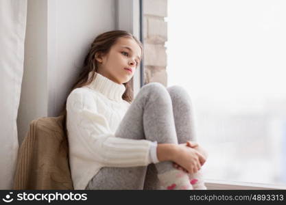 childhood, sadness and people concept - sad beautiful girl in sweater sitting on sill at home window in winter