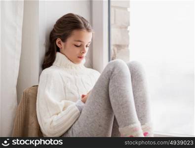 childhood, sadness and people concept - sad beautiful girl in sweater sitting on sill at home window in winter. sad girl sitting on sill at home window in winter